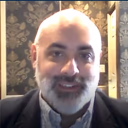 Smiling bald man with a gray beard wearing a dark jacket and light shirt in front of a patterned wallpaper.