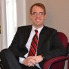 Man wearing glasses, a black suit, white shirt, and red-striped tie, sitting in a chair and smiling.